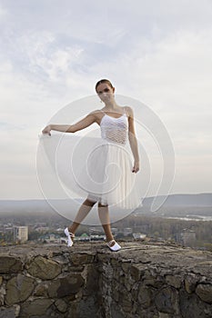 Young ballerina in white dress and satin ballet shoes posing on the edge of old fortress wall on a grey sky background