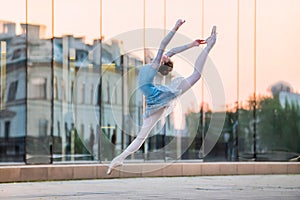 Young ballerina in a white leotard is dancing on pointe shoes against the backdrop of the reflection of sunset in the city, frozen