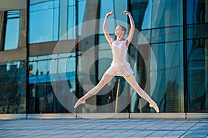 Young ballerina in a white leotard is dancing on pointe shoes against the background of a cityscape, frozen in jump