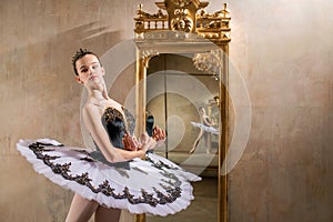 Young ballerina in a white tutu poses near beautiful old mummer in vintage interior, reflecting many times