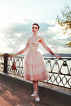 a young ballet dancer stands in a pink dress and Czechs with ribbons on a cast-iron railing of the river fence on a warm photo
