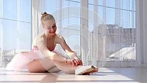 Young ballet dancer in tutu sitting on the floor and tying pointe shoes