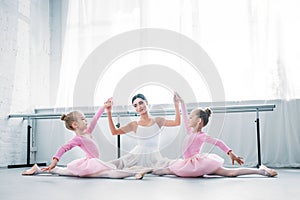 young ballet teacher with children in tutu skirts sitting and holding hands while exercising