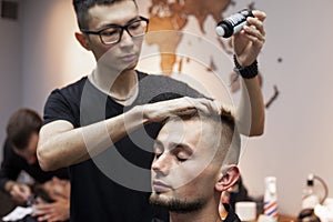 Young barber kazakh works in a barbershop, handsome guy does hair styling for a client with dry gel in a hairdresser