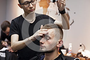 Young barber kazakh works in a barbershop, handsome guy does hair styling for a client with dry gel in a hairdresser