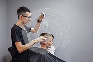 Young barber kazakh works in a barbershop, a handsome guy makes hair styling to a client with dry gel on a gray wall background
