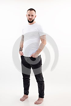 Young barefoot bearded man with short dark hair in white T-shirt, black jeans, standing, thrusting hands into pockets.