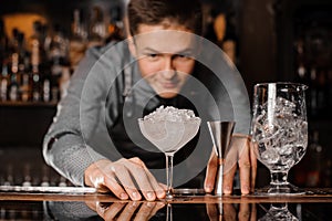 Young barman looking at the cocktail glass filled with ice
