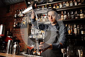 Young barman pouring alcoholic drink from one metal glass into another