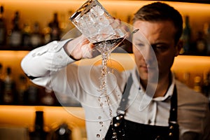 Young barman pouring water from a glass with ice cubes