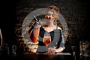 Young bartender girl attentively pours carbonated drink to glass with negroni cocktail.