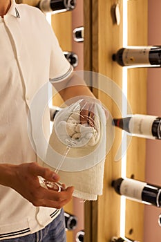 A young bartender is wiping towel wine glasses at work in the restaurant on the background of bottles of wine