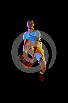 Young basketball athlete training dribbling technique in motion against black studio background in mixed neon light.