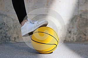 Young basketball player treading with her white shoe a yellow basketball in an urban court