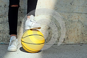 Young basketball player treading with her white shoe a yellow basketball in an urban court