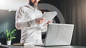 Young bearded atrractive businessman in white shirt is standing near desk in front of laptop, reading documents.