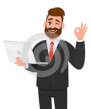 Young bearded business man is holding/showing a latest new laptop and gesturing/making okay or OK sign with hand fingers.