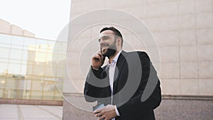Young bearded business man talking on cell phone making deals with cup of coffee near modern office buildings