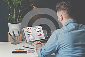 Young bearded businessman in denim shirt is sitting in office at table and is using laptop with charts, graphs