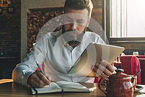 Young bearded businessman sits in cafe, home at table, holding tablet computer and writes in notebook.