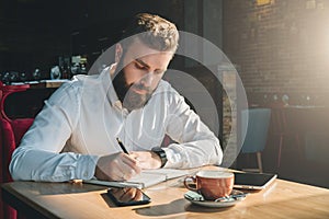 Young bearded businessman sits in cafe, home at table and writes in notebook. On table tablet computer, smartphone.