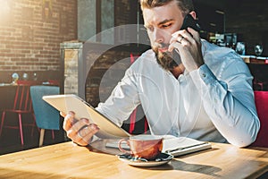 Young bearded businessman sits in cafe at table, talking on mobile phone, holding tablet computer.Man is working