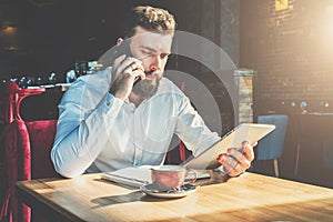 Young bearded businessman sits in cafe at table, talking on mobile phone, holding tablet computer.Man is working