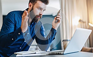 Young bearded businessman sits in front of computer and looks at laptop screen with astonishment, raising his hands up.