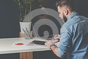 Young bearded businessman sits in office at table,using digital tablet and explores charts,making notes.