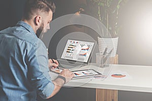 Young bearded businessman sits in office at table,using tablet computer and explores charts,making notes.