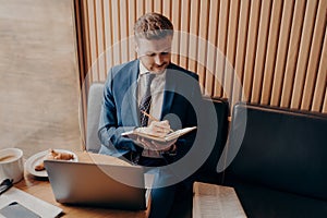 Young bearded businessman sitting in cafe and writing down notes during online webinar