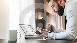 Young bearded businessman sitting at desk in front of laptop with graphis, charts, diagrams on screen