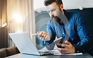 Young bearded businessman is sitting in front of computer, holding smartphone and showing pen on laptop screen.