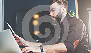 Young bearded businessman sitting in office at table, reading documents and working on laptop. Man blogging, chatting