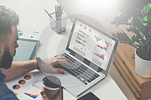 Young bearded businessman is sitting in office at table, typing on laptop with graphs,diagrams and charts on screen