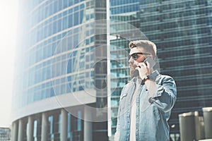 Young bearded businessman in sunglasses and shirt stands on city street and talks on his cell phone.Summer sunny day