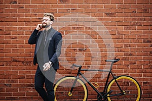 Young bearded Businessman talking on smartphone commuter with bicycle in city street against brick wall