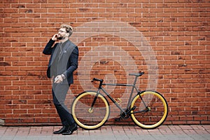 Young bearded Businessman talking on smartphone commuter with bicycle walking in city against brick wall