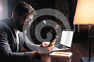 Young bearded businessman using phone while sitting by the wooden table in modern office at night. People working mobile devices