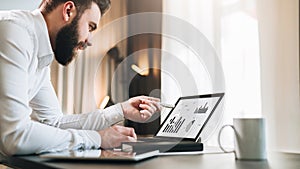 Young bearded businessman in white shirt is sitting at table in front of computer, pointing with pen on graphs, charts