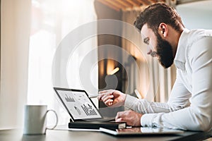 Young bearded businessman in white shirt is sitting at table in front of computer, pointing with pen on graphs, charts