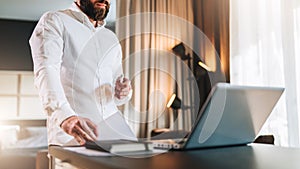 Young bearded businessman in white shirt is standing near desk in front of laptop, holding documents. Freelancer working