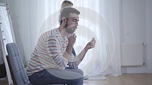 Young bearded Caucasian man looking at wedding ring in hand while nervous wife walking at the background. Stressed