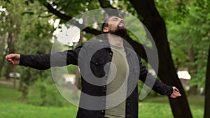 Young bearded caucasian man stands in a park between trees and hands up enjoying nature