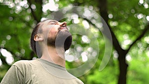 Young bearded caucasian man stands in a park between trees and head up enjoying nature. close up