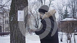 Young bearded Caucasian man talking on the phone and looking at missing person ad hanging on the tree. Empathic guy in