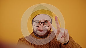 Young bearded Caucasian man talks, smiles, shows thumb up and sign of freedom and peace, points finger to the camera