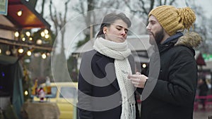 Young bearded Caucasian man in yellow hat using smartphone as his girlfriend or wife talking with him having ironical