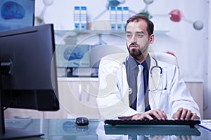 Young bearded doctor with both hands on the keyboard typing the recipe for a patient