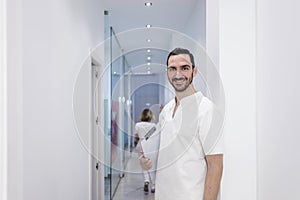 Portrait of a Young smiling bearded doctor holding a folder while leaning on white wall and looking to camera at a clinic corridor